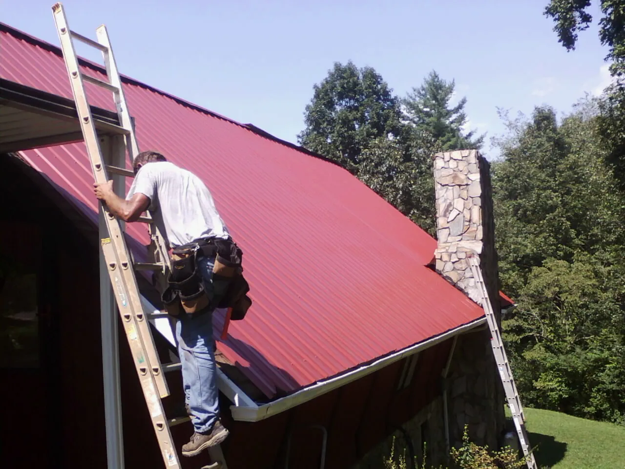 Roofer on a ladder beside a red metal roof and stone chimney — Soffit & Fascia Repair by Roofing Brothers in Brownsburg
